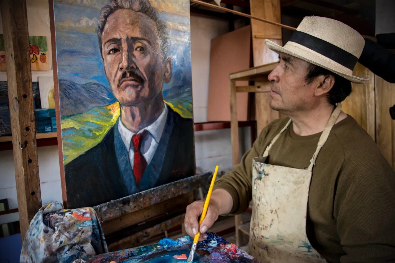 Alejandro Galindo Yauris, con sombrero y mandil, pinta en su taller ante un retrato al óleo