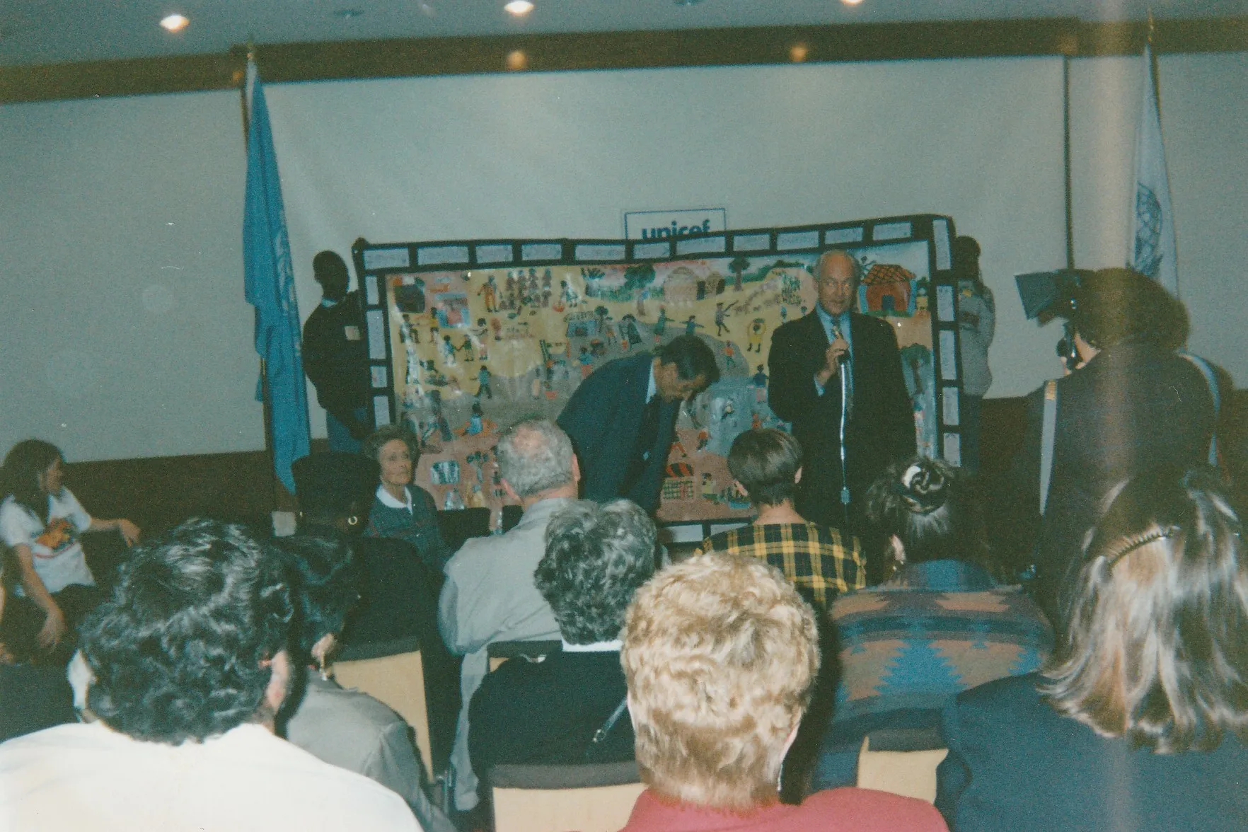 Un acto en una sala con la bandera de la ONU y el rótulo de UNICEF, con un gran panel pintado de escenas de aldea en el escenario