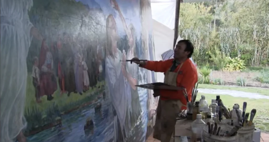 Alejandro Galindo, con chaqueta naranja y mandil, pinta con la paleta en la mano una escena de gran formato con una multitud a la orilla de un río, al aire libre bajo un toldo