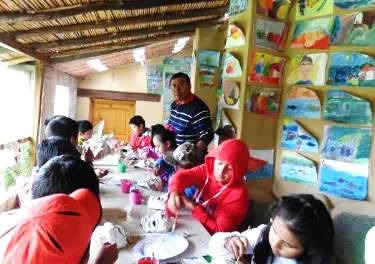 Niños pintan en una mesa larga bajo un techo de cañas, con sus dibujos colgados en la pared